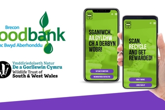 A graphic with white background showing with Brecon Foodbank and The Wildlife Trust of South & West Wales' logo. On the right there a person holding phone showing a green screen the scan, recycle, reward app.