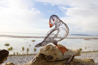 A puffin sculpture made of wire, perched on a piece of driftwood in front of the coast. 