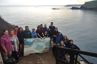 A group of young people stand on the jetty steps. They are smiling and posing with a large painting of the island.