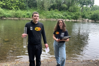 Two people stand by the riverside, holding water monitoring equipment and a clipboard