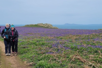 Karen and friend on a path on Skomer Island on a sunny spring day. There are surrounded by bluebells and the sea is the in the background.