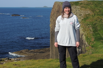 Lizzie standing in front of a coastal cliff - the sea is deep blue. 