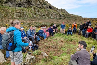 A photo of the group on a guided walk. They are sat around the edge of an Iron Age roundhouse.
