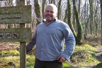 Gareth next to a wooden sign in a woodland.