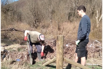 Two volunteers stood either side of a fence that they are repairing. One volunteer is holding a shovel.