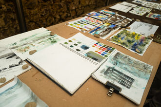A table is spread out with sketchpads, colour charts and painting tools.