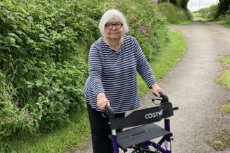 A woman stood with a walking aid on a path with rich green verges on each side. 