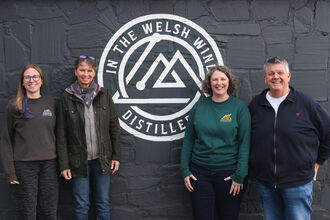 Fundraisers and In the Welsh Wind team standing in front of a grey wall with In the Welsh Wind logo.
