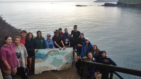 A group of young people stand on the jetty steps. They are smiling and posing with a large painting of the island.
