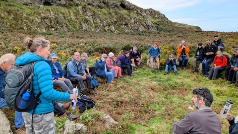 A photo of the group on a guided walk. They are sat around the edge of an Iron Age roundhouse.
