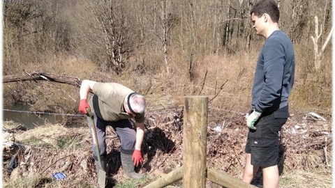 Two volunteers stood either side of a fence that they are repairing. One volunteer is holding a shovel.