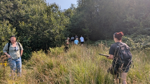 A group of four people in the background walk away from the camera toward the treeline. Two people stand in the foreground in a sunny field. The person on the right of the image holds a clipboard.