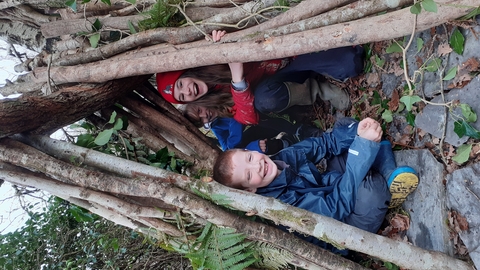 3 children sitting in a woodland  shelter made from branches