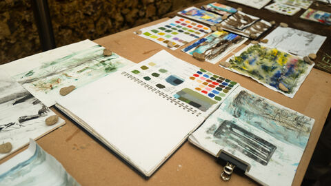 A table is spread out with sketchpads, colour charts and painting tools.