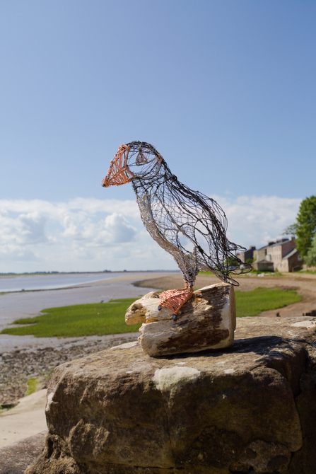 A puffin sculpture made of wire, perched on a piece of driftwood in front of the coast. 