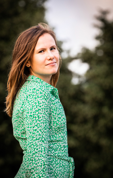 Zoe stands side on, looking back towards the camera. She is wearing a green dress with a white and black spotted pattern