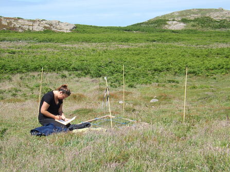A woman kneeling in a grassy landscape writing on a clipboard
