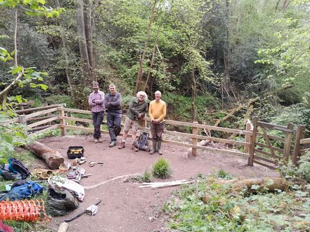 A group of people stood in front of a new wooden fence with large trees around them.