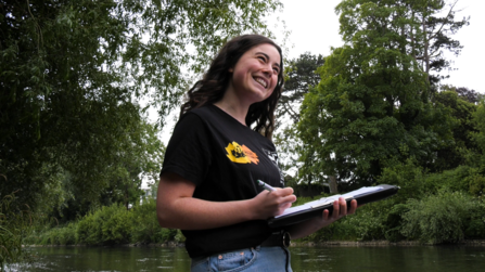 A smiling woman holds a clipboard and pen while standing by the riverside. She conducts a pollution survey as part of the Save our Taff campaign.
