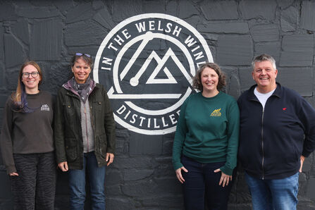Fundraisers and In the Welsh Wind team standing in front of a grey wall with In the Welsh Wind logo.