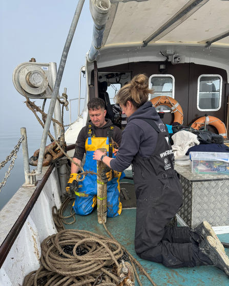 Two people kneeling on a boat at sea. They are next to a large pile of rope and holding a cylindrical device covered in seaweed and mud. 