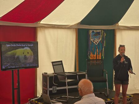 A woman giving a talk in a marquee. She is stood next to a screen with a picture of a seal on it.