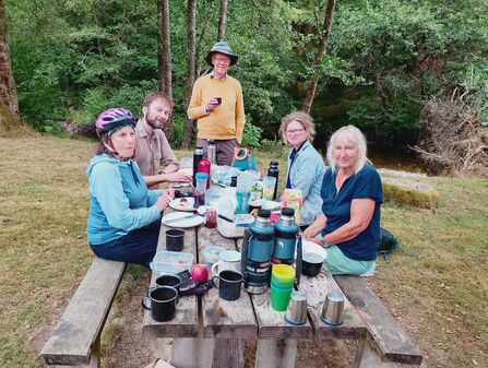 A group of people sat on a picnic bench and sharing food. They are smiling at the camera.