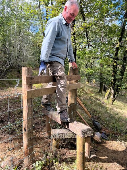 A man walking over a newly-built stile in a fence. There are tools laying in the background.